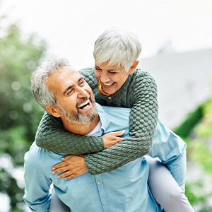 Smiling woman in green sweater riding laughing man’s back outside near green trees