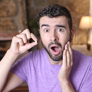 Man in purple t-shirt looking shocked holding tooth in one hand and his jaw with the other